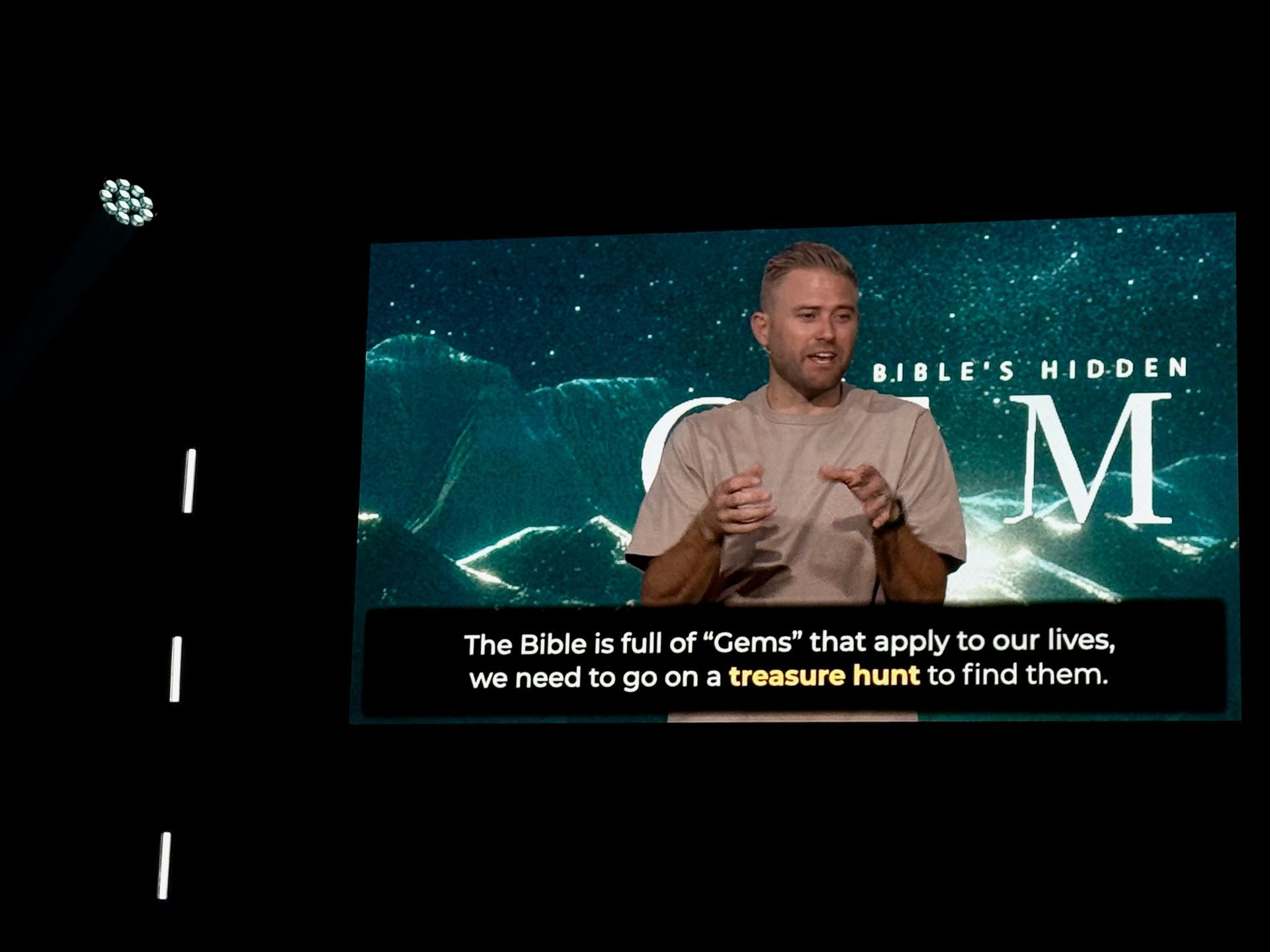 A man gestures while speaking in front of a screen with text 
