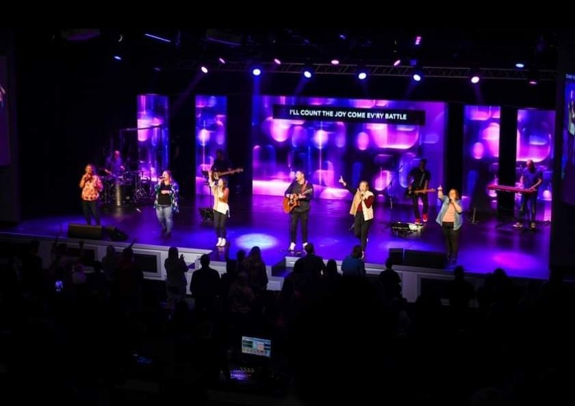 A worship band performs on a stage with purple lighting. People sing, play instruments, and worship.