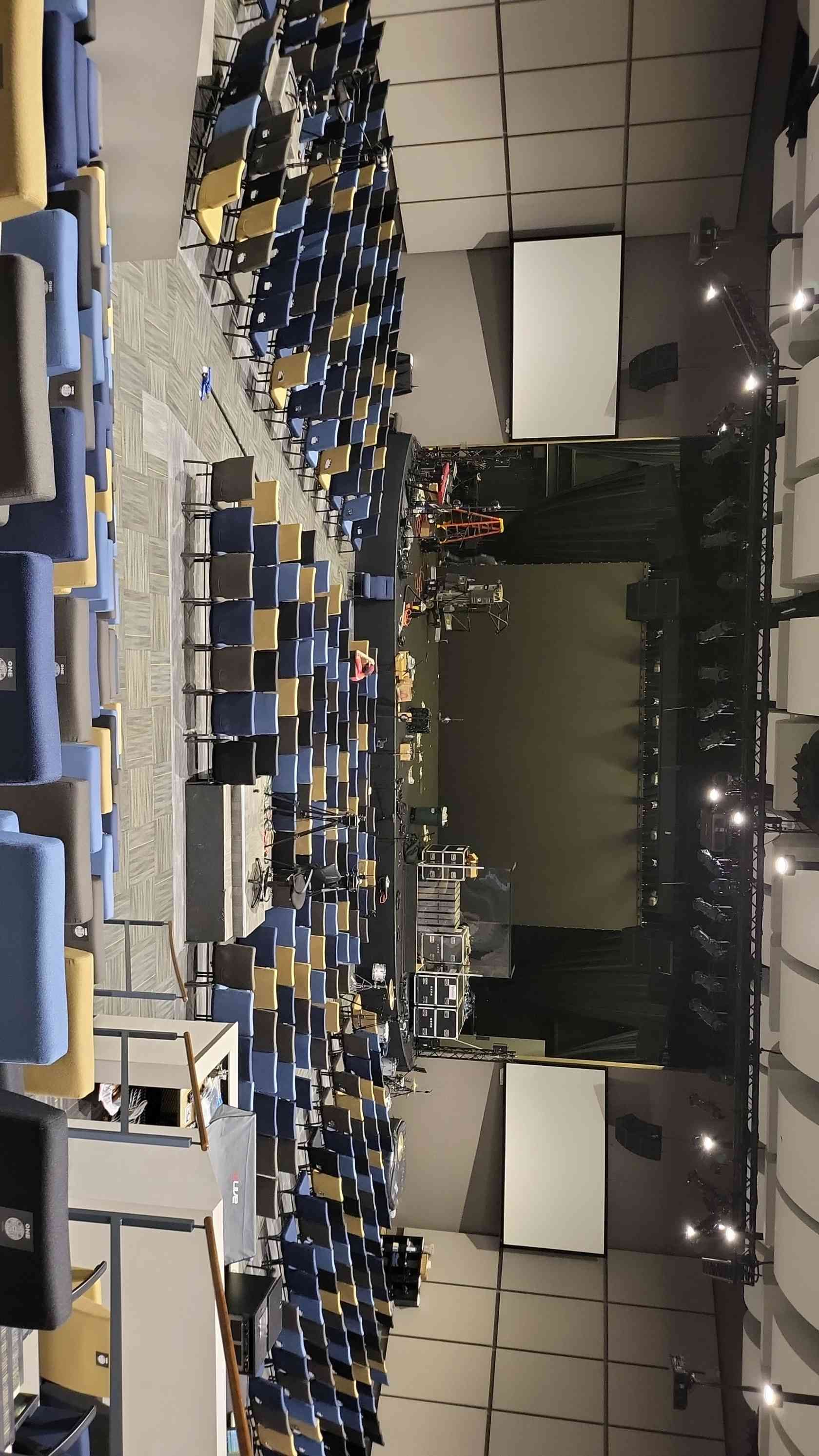 Auditorium with blue seats, stage, and two white screens. Dim lighting with a few lights on.