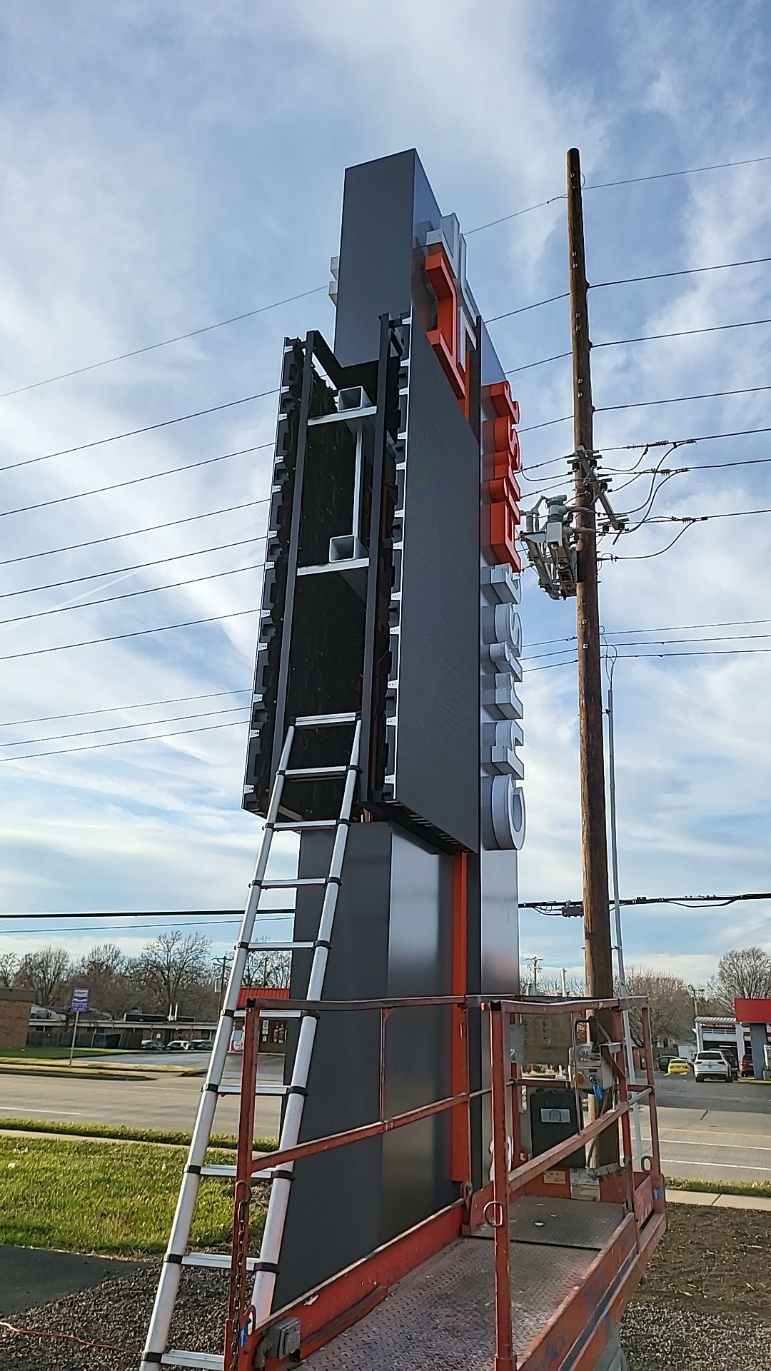 Construction workers on a tall, grey and orange sign, with a ladder. A utility pole is nearby.