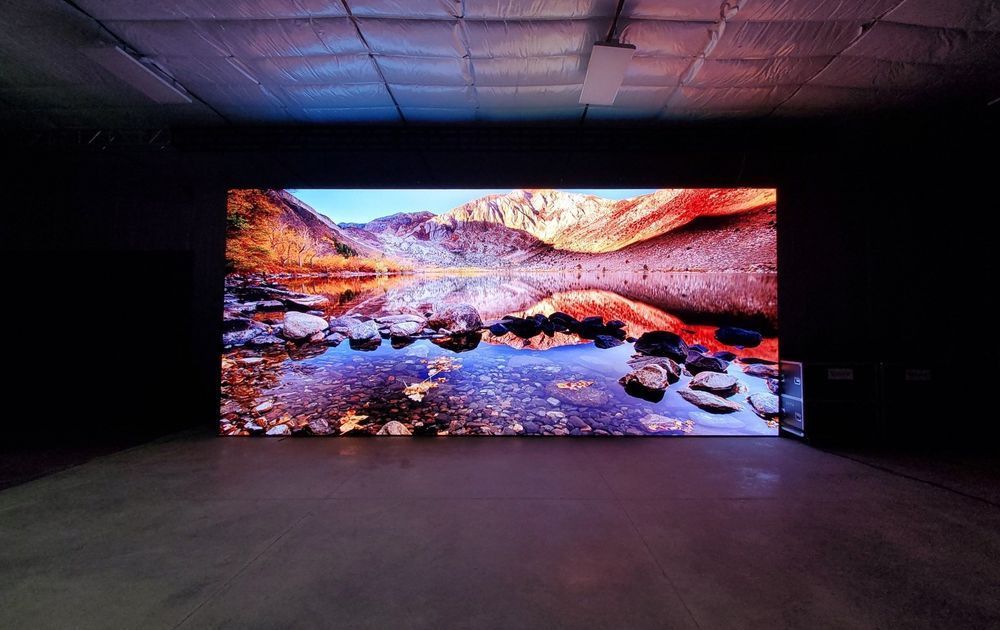 Large, rectangular digital screen displaying a mountain lake scene; a dark room.