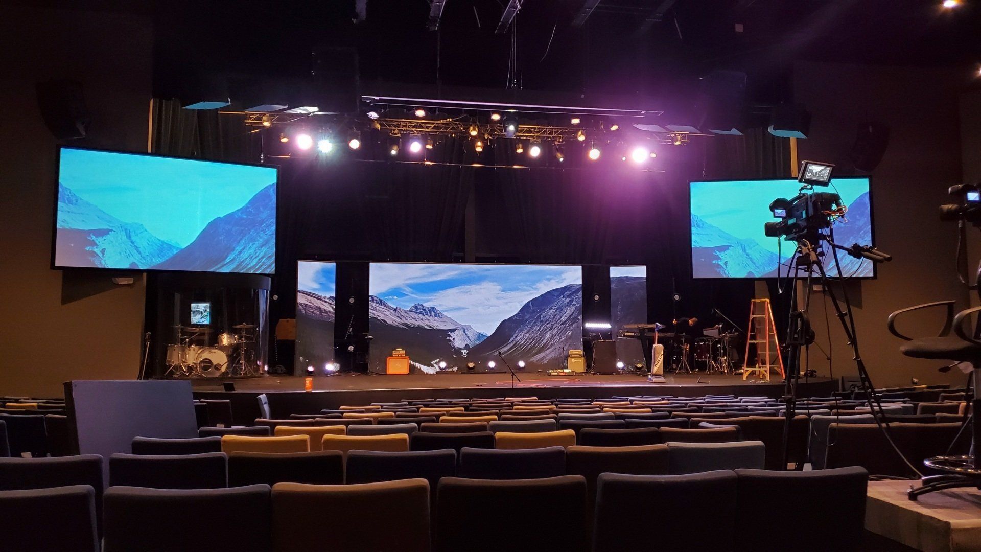 Auditorium with stage, screens, and camera equipment, featuring a mountain landscape. Rows of seats are visible.