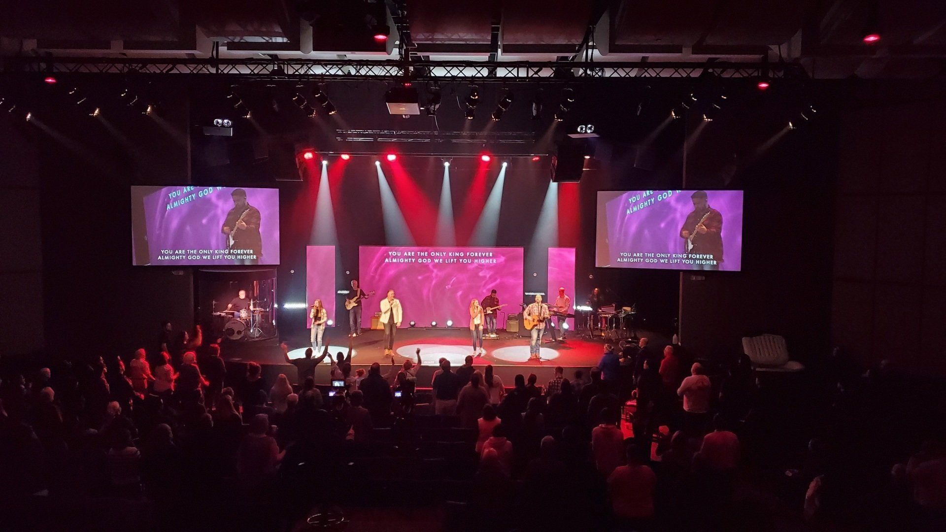 A live worship service with a band on stage, singing with a crowd in a dark auditorium. Red lighting.
