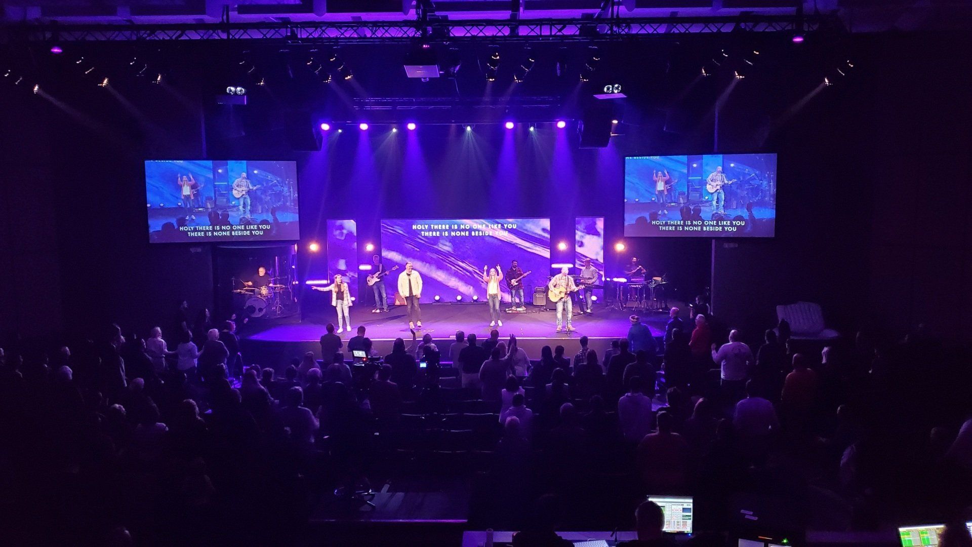 A band performs on stage under purple lights at a church service; a crowd watches from below.