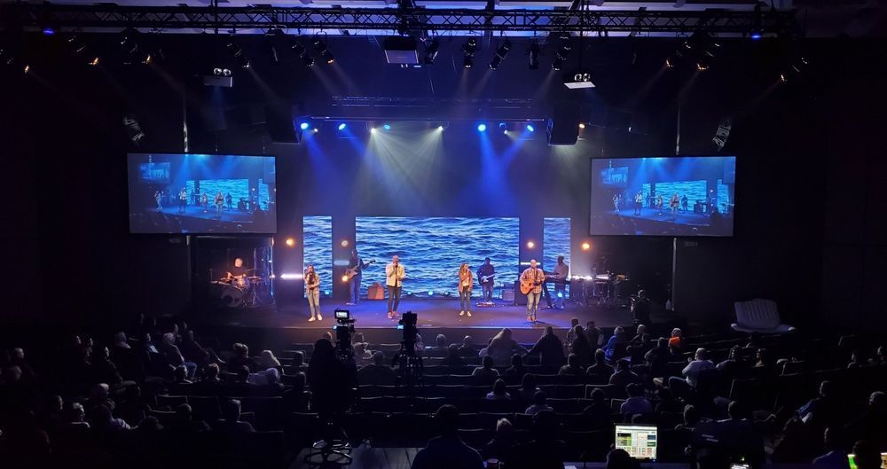 Band performing on a stage with screens displaying images, in front of an audience, lit by blue lights.