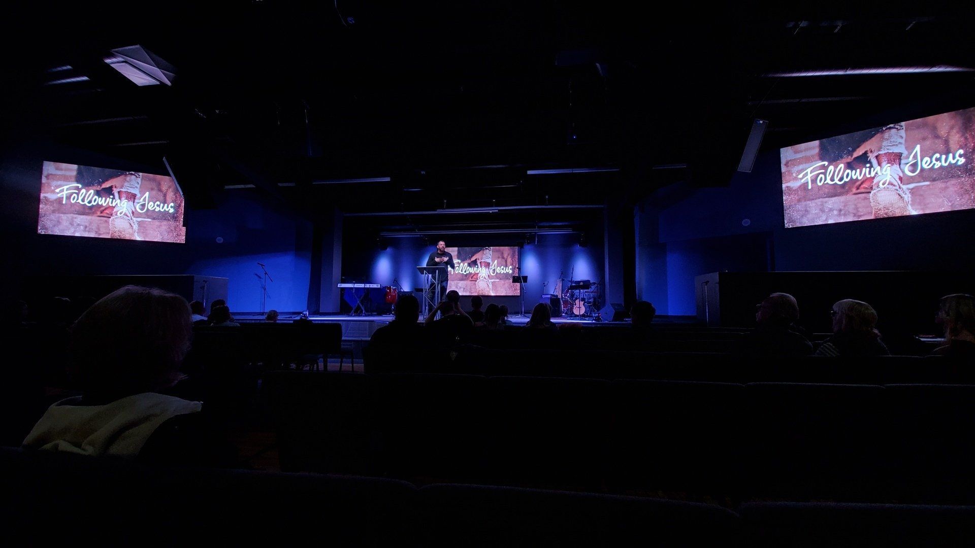 A man performs on a stage in a dark auditorium. Two screens display text, blue lighting. Audience in the foreground.