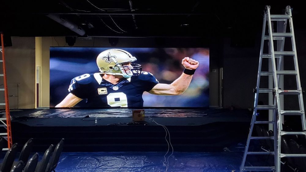 Drew Brees celebrating on a large screen, with a ladder to the right in a dark setting.