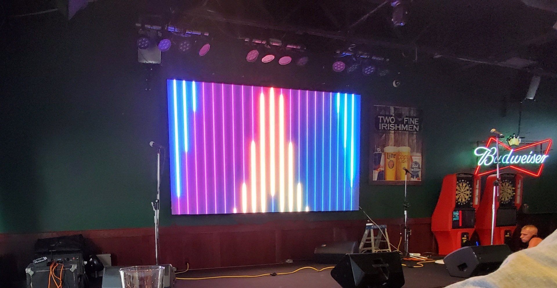 Stage with a colorful LED screen displaying vertical stripes, Budweiser sign in a bar.
