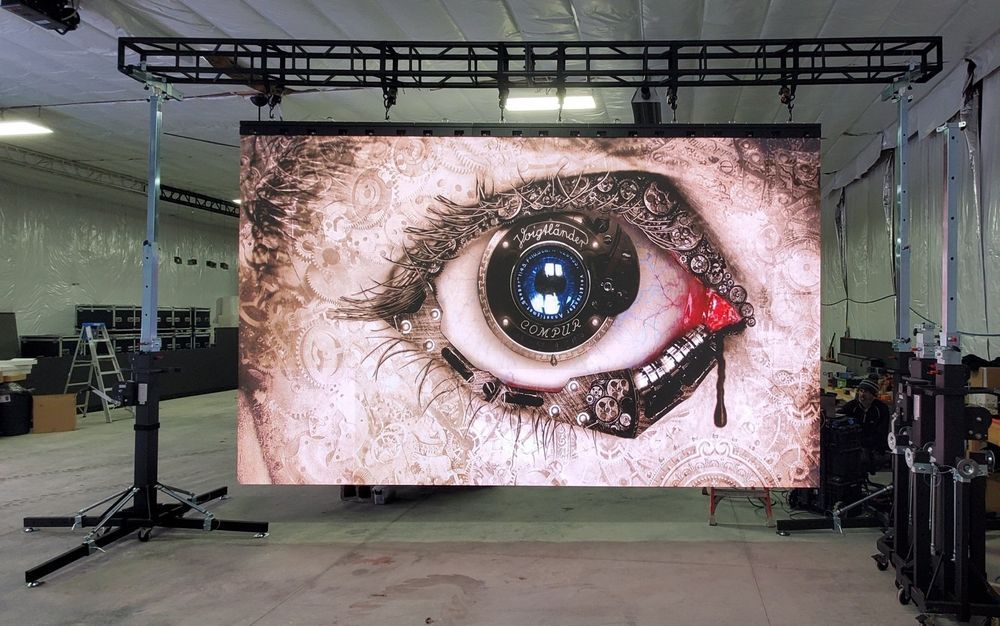Large video screen displays a close-up of an eye with mechanical parts in a warehouse. Screen is held by a truss structure.