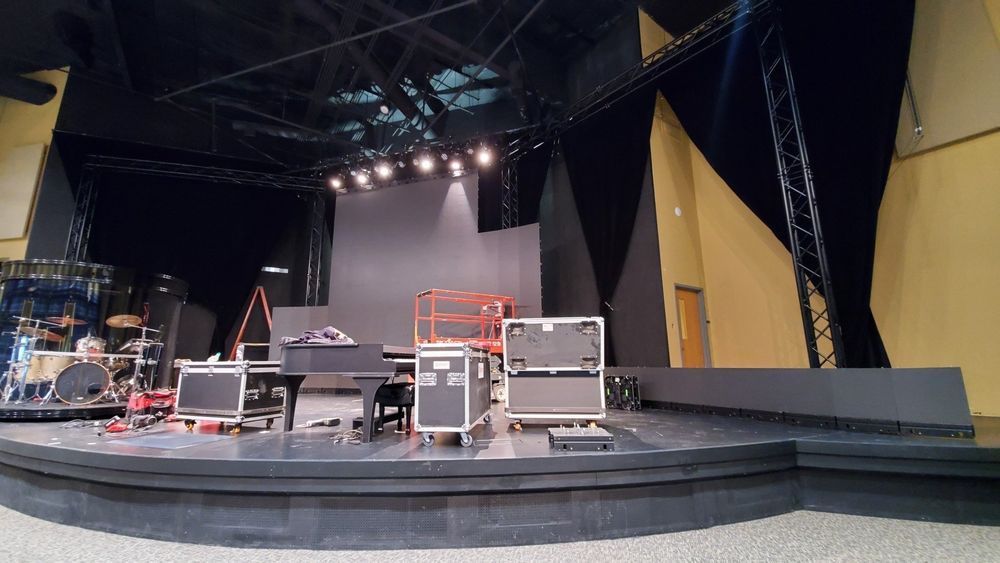 Stage set with musical equipment: drums, piano, speakers. Black, gray and yellow backdrop.