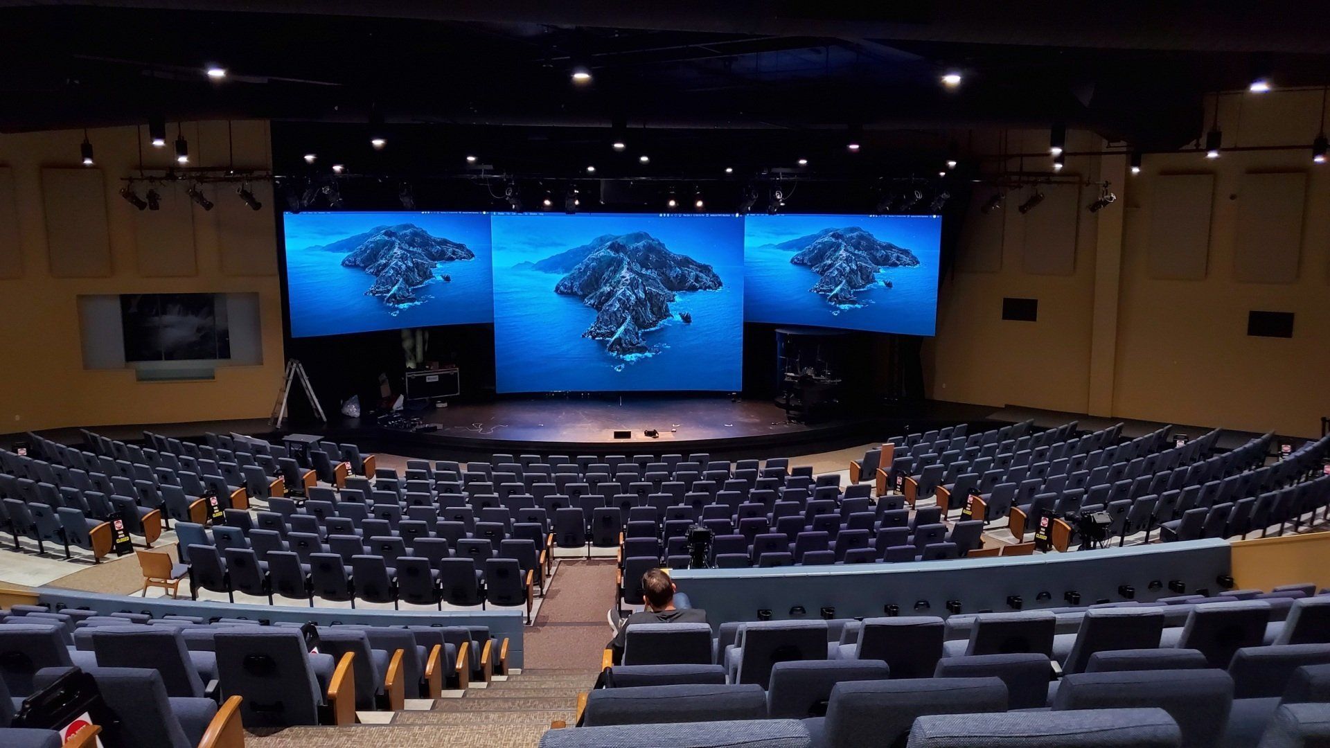 Auditorium with rows of blue chairs facing a stage with three screens displaying a blue mountain image.