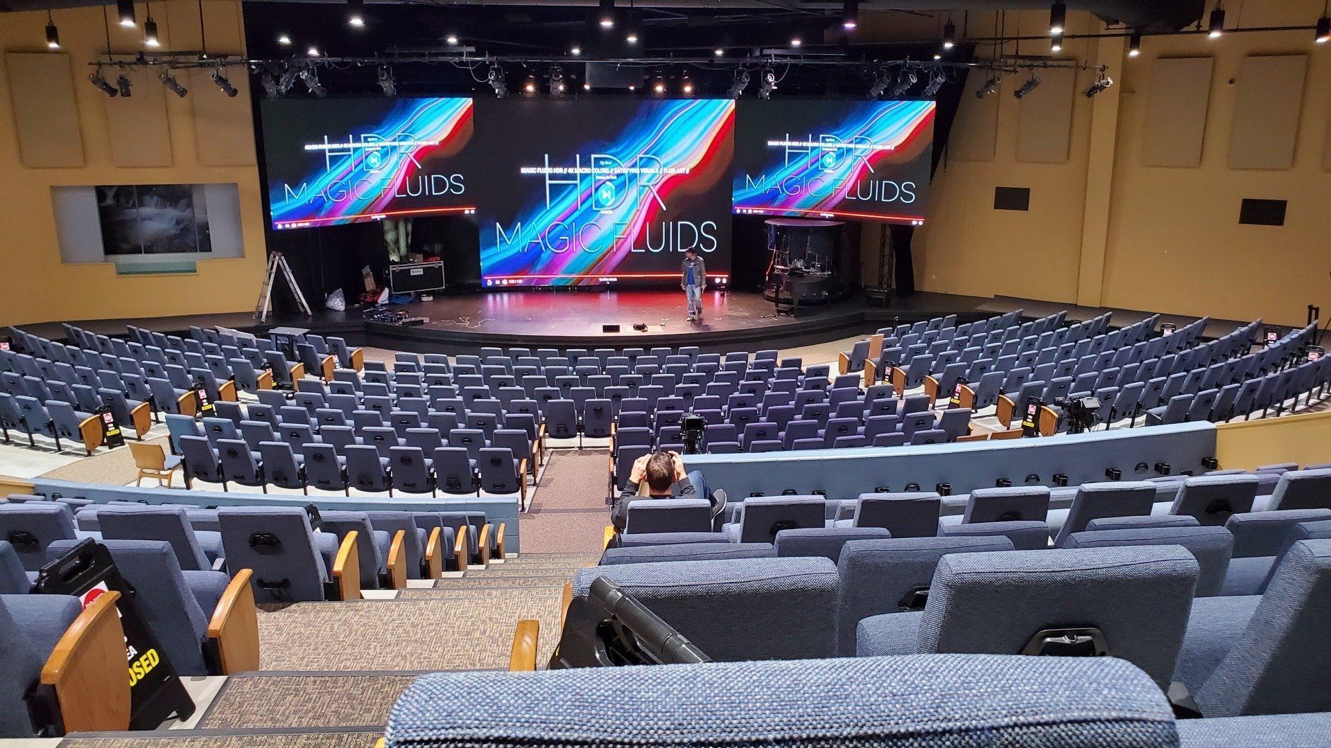 Auditorium with rows of blue seating facing a stage with a large video display.
