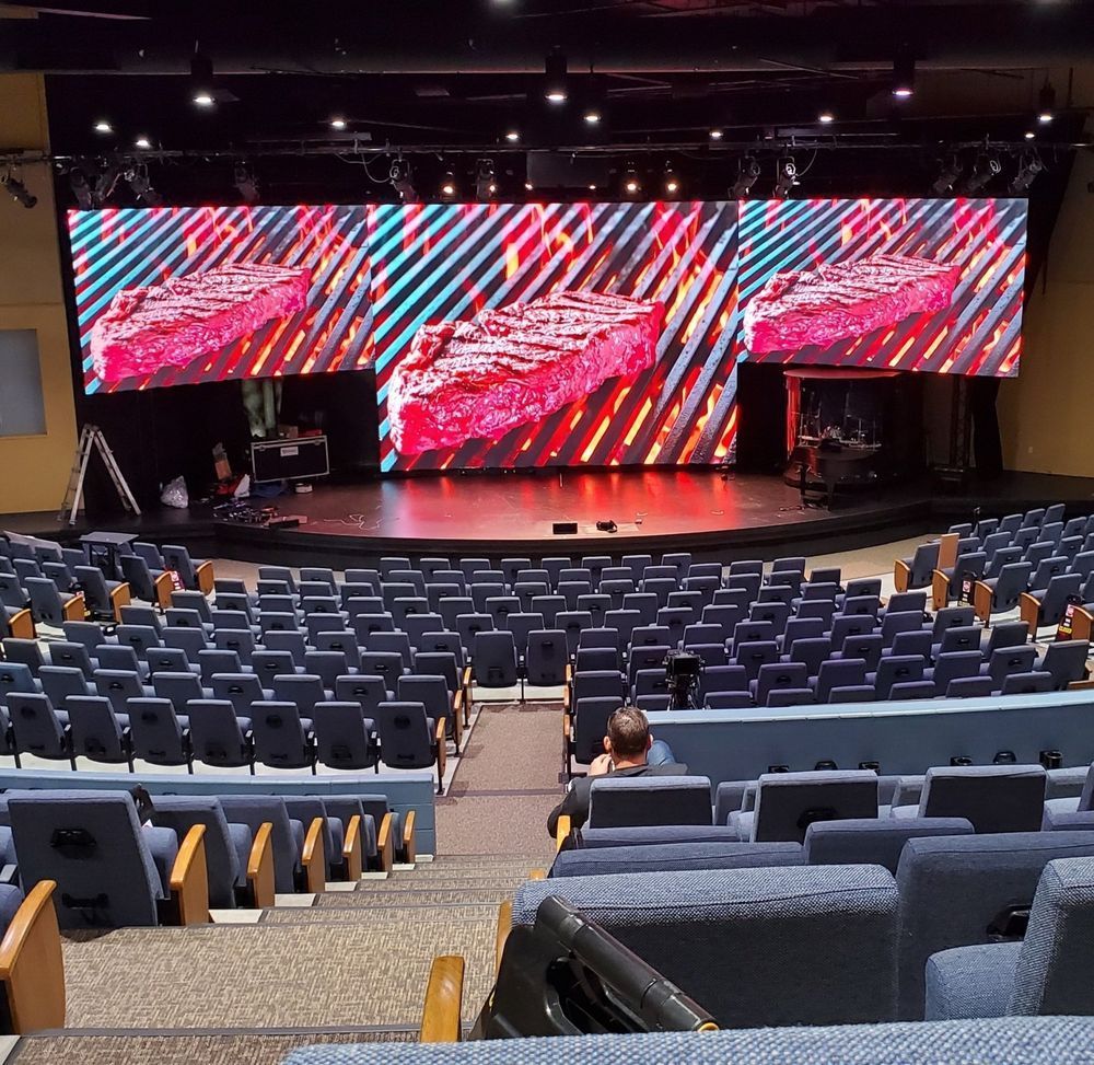 Auditorium with rows of blue seats facing a stage with three screens displaying a steak.