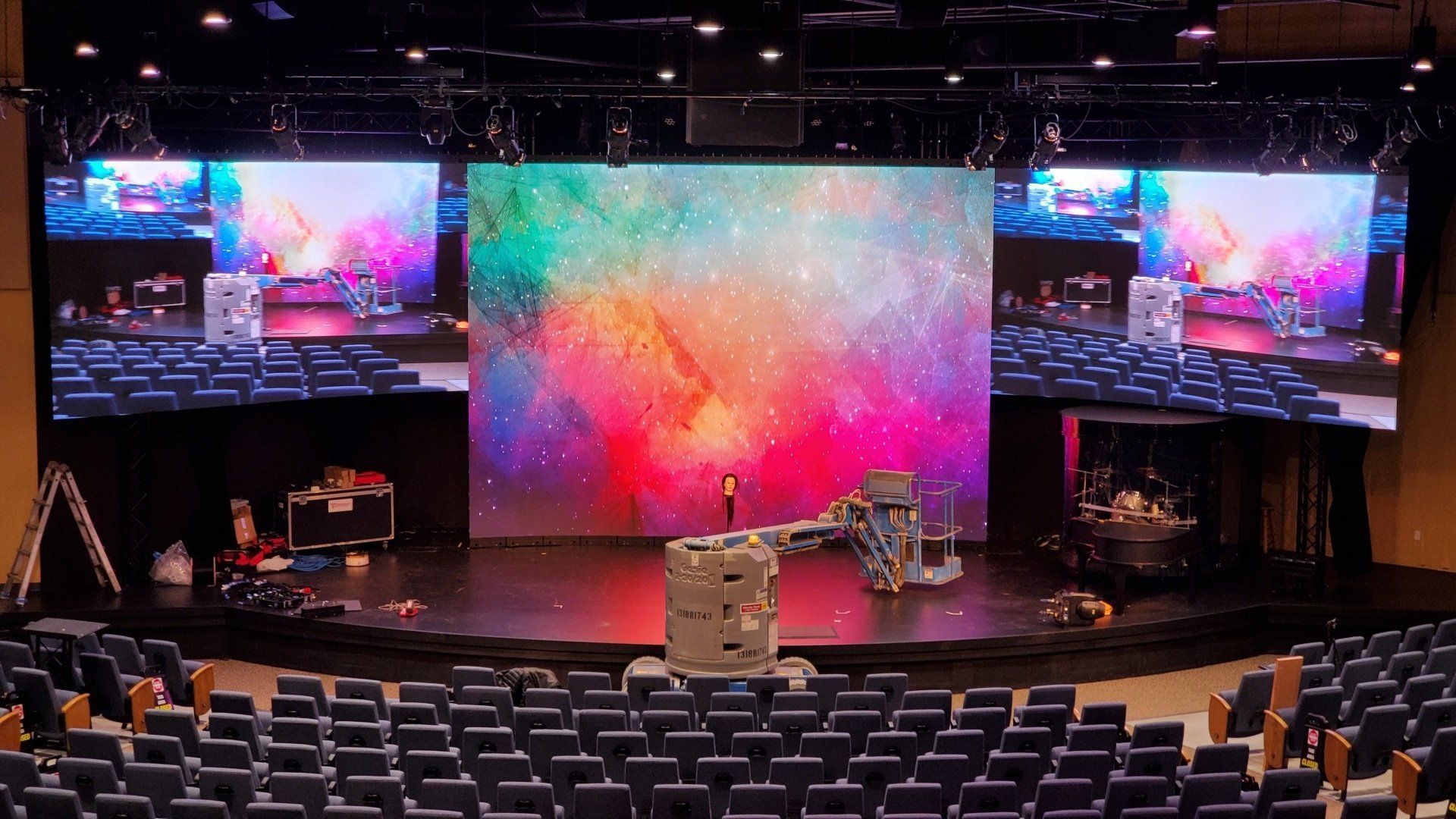 Stage with large LED screens displaying a colorful galaxy. A speaker stand stands in front. Rows of seating are in foreground.
