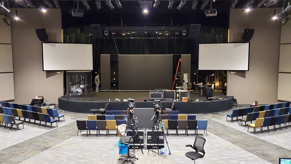 A modern church stage with screens, sound equipment, and seating. Preparations underway.