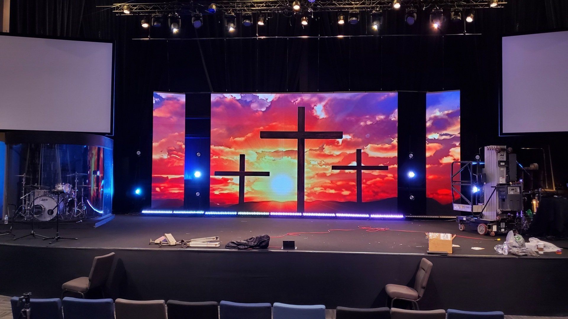 Stage with three crosses silhouetted against a sunset backdrop, drum set left, and equipment right.