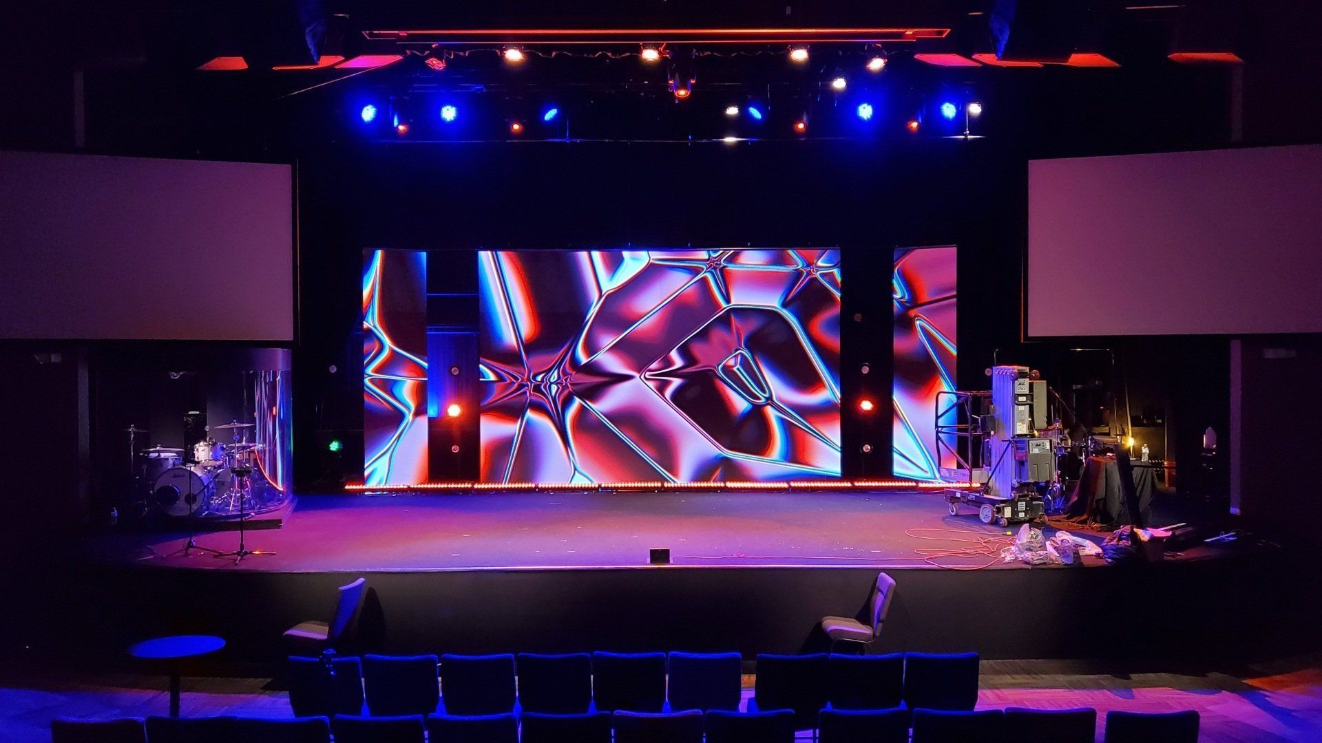 Stage with a large screen displaying a geometric pattern, flanked by screens. Empty chairs in foreground. Stage lights.