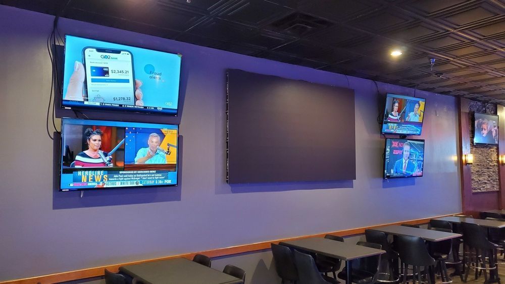 Wall with multiple TVs displaying various content in a restaurant setting. Purple wall and dark furniture.