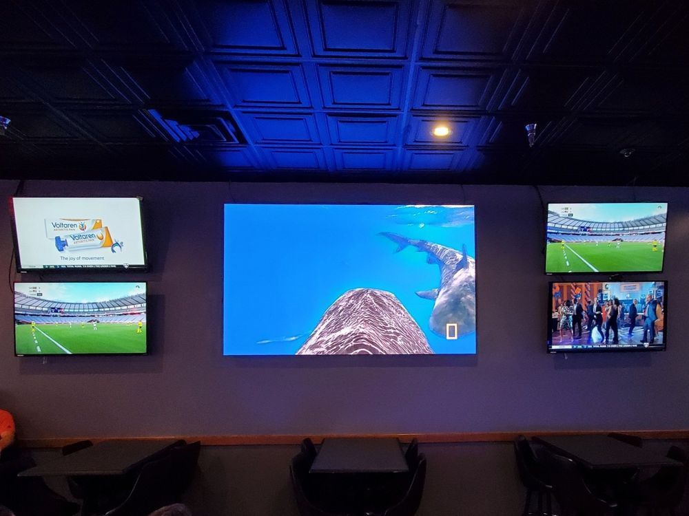 A wall with six TVs displaying content. The center screen shows an underwater view, while others show sports and graphics.