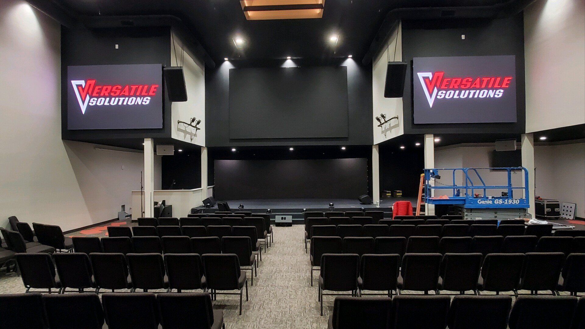 Auditorium with rows of black chairs facing a stage with two screens displaying a logo.