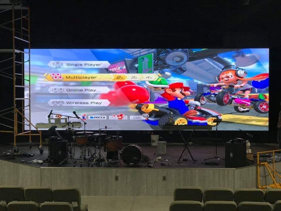 Mario Kart game menu displayed on a large screen on a stage with a drum set and seating.