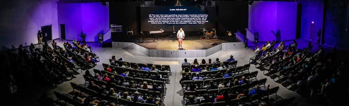 A person standing on a stage addressing an audience seated in a theater-like venue. Purple lighting.