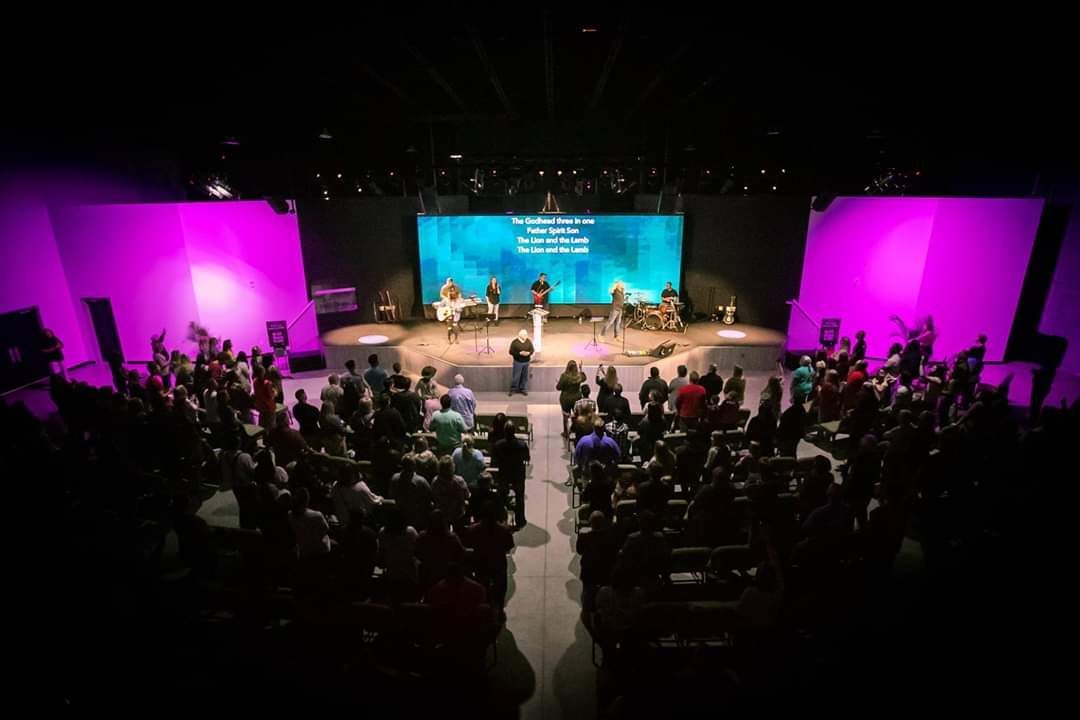 Audience watching a band perform on stage with a large screen backdrop. Pink lighting and purple walls.