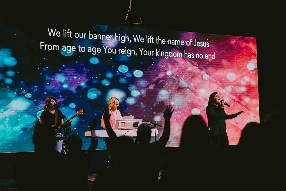 Worship service with musicians on stage; lyrics displayed on a screen with colorful background; audience with raised hands.