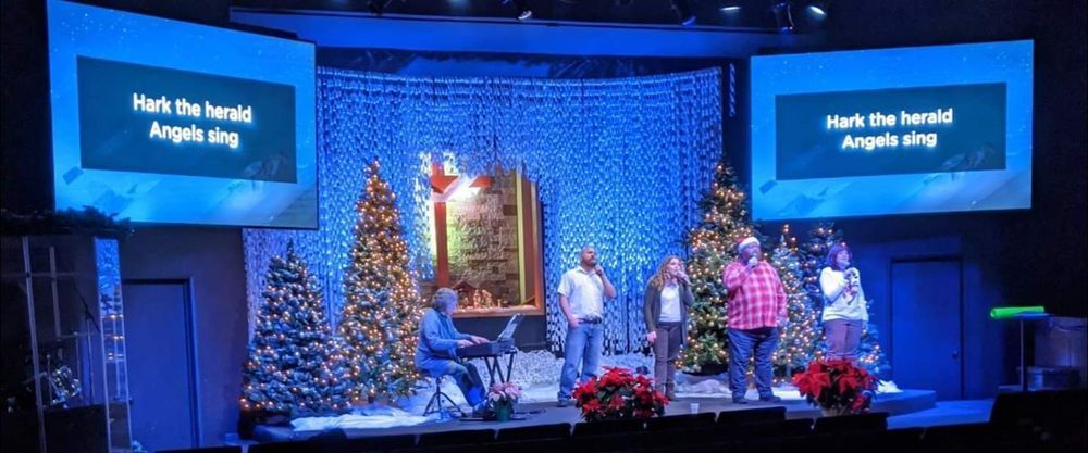 A stage with a group singing, Christmas decorations, and screens displaying lyrics: 
