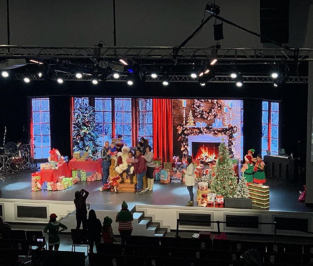 Santa Claus on stage surrounded by elves, children, and gifts. Festive scene with a Christmas backdrop.