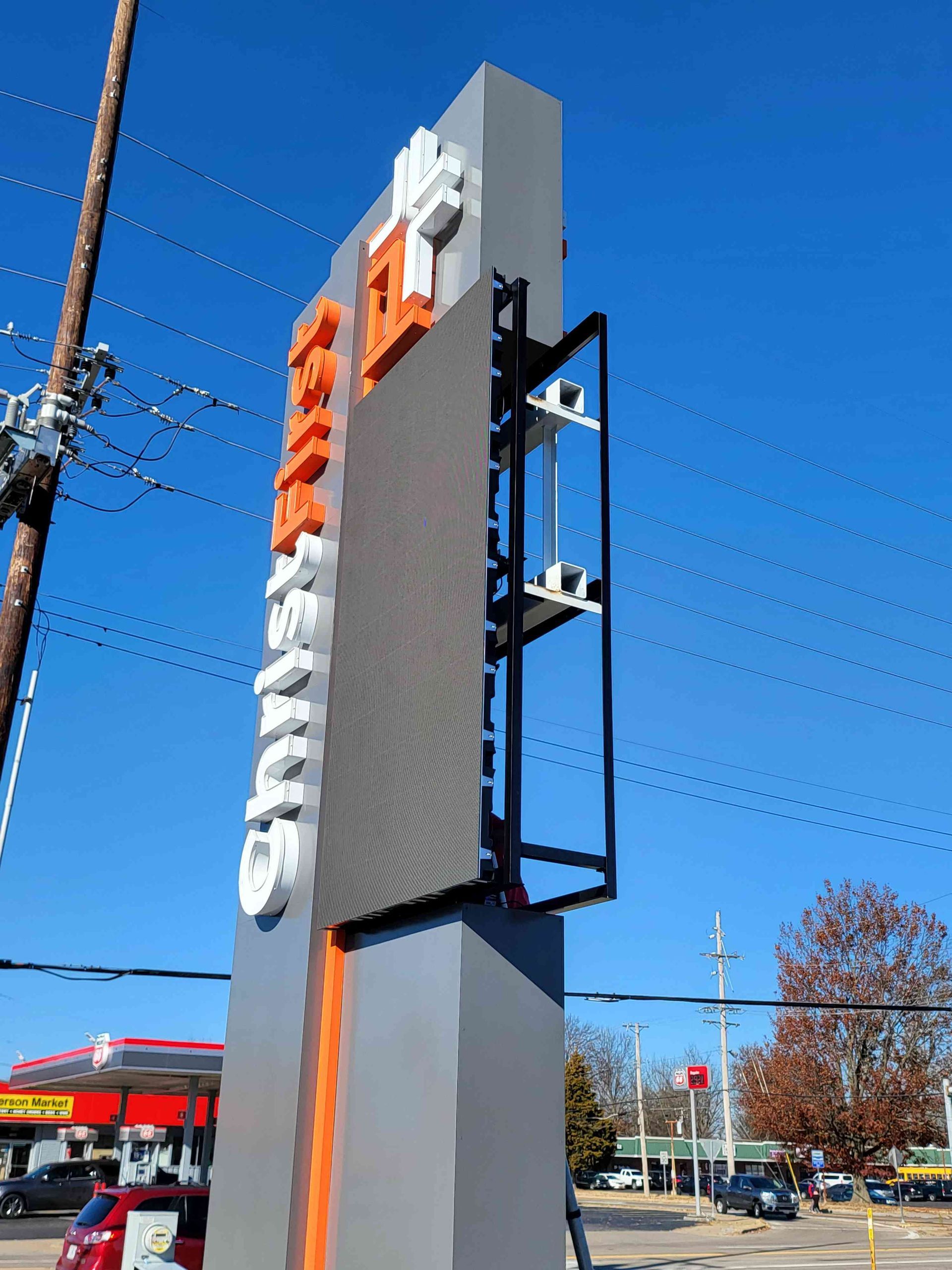 Tall grey and orange sign with LED display next to a gas station.