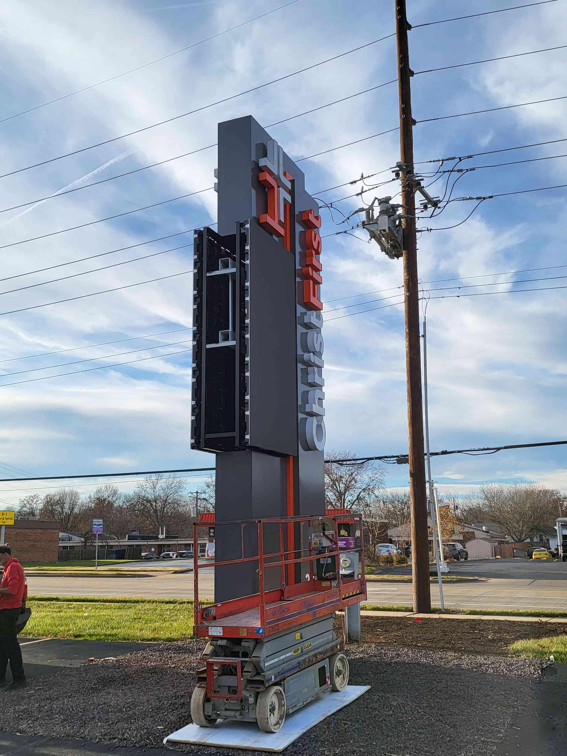 Construction worker on a lift installing a digital sign for a business. Gray and red colors. Outdoors with power lines and trees.