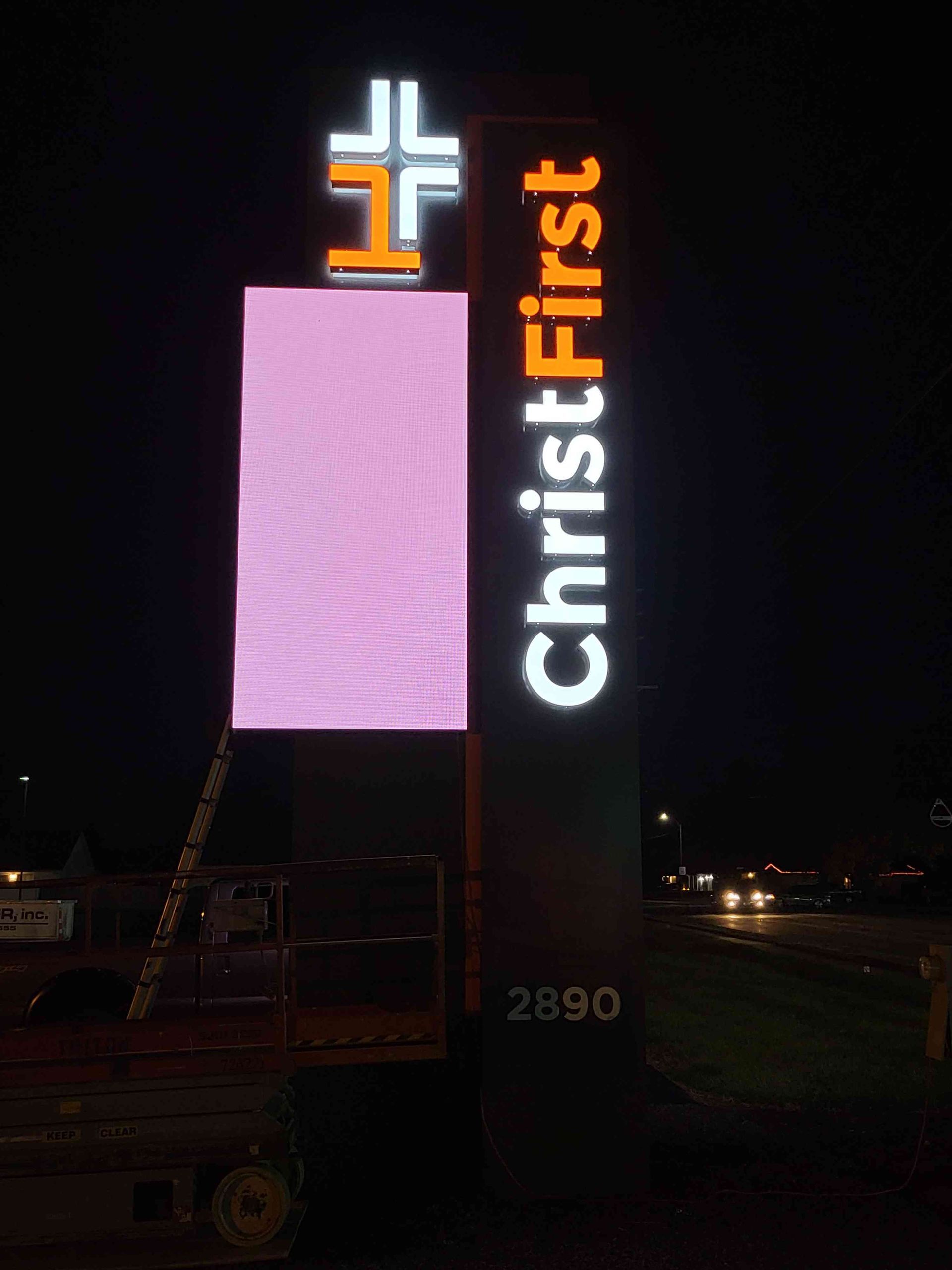 Christ First church sign at night; lighted cross on top, digital screen lit pink, black and orange tower, address 2890.