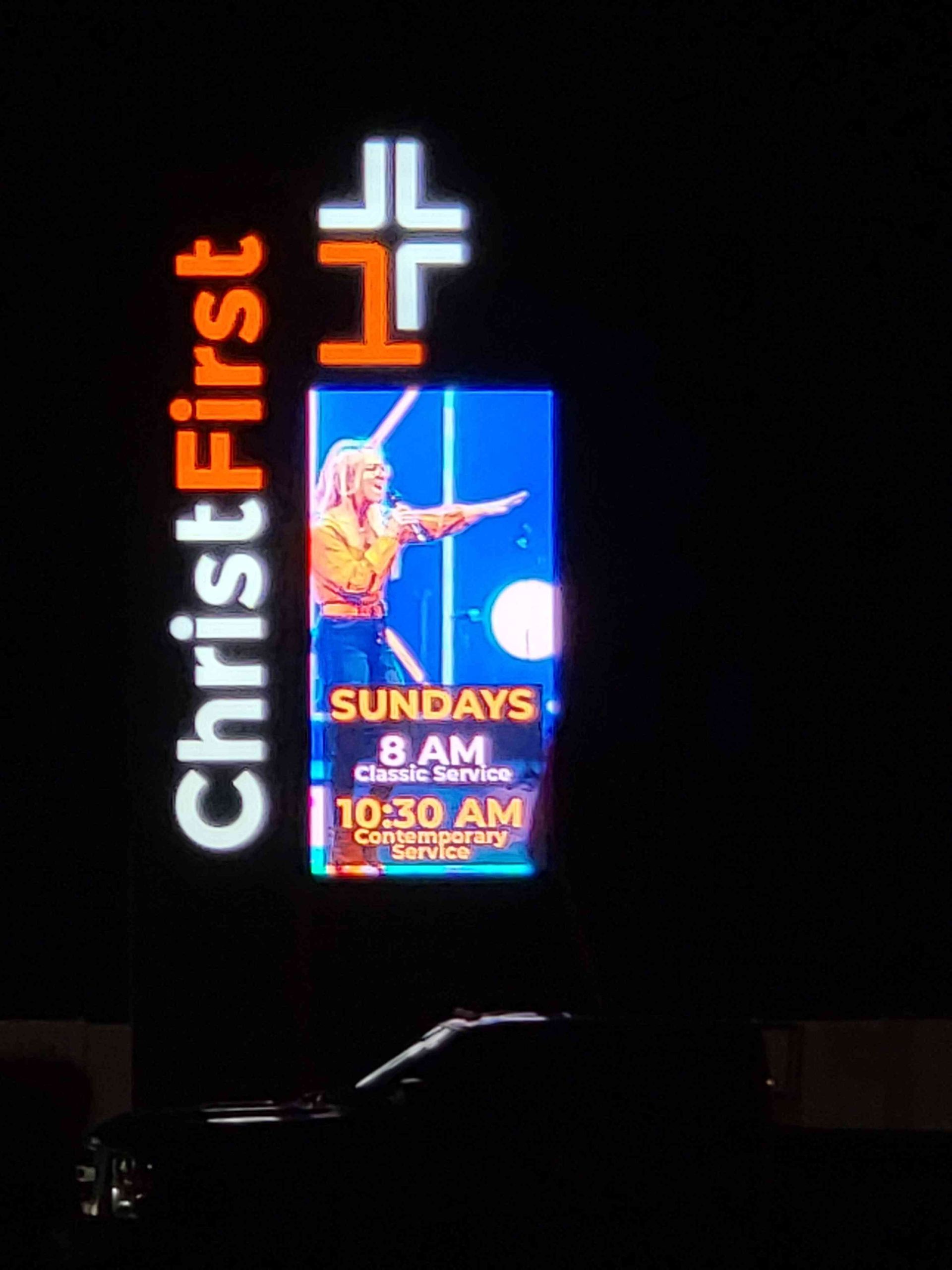 Christ First Church sign at night: Features service times, speaker image, orange/white cross logo, and 