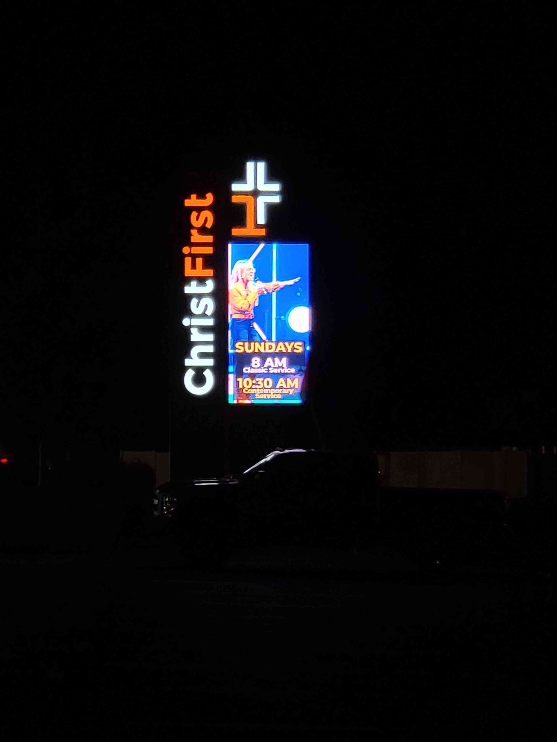 Sign for Christ First church at night with a cross logo and service times.