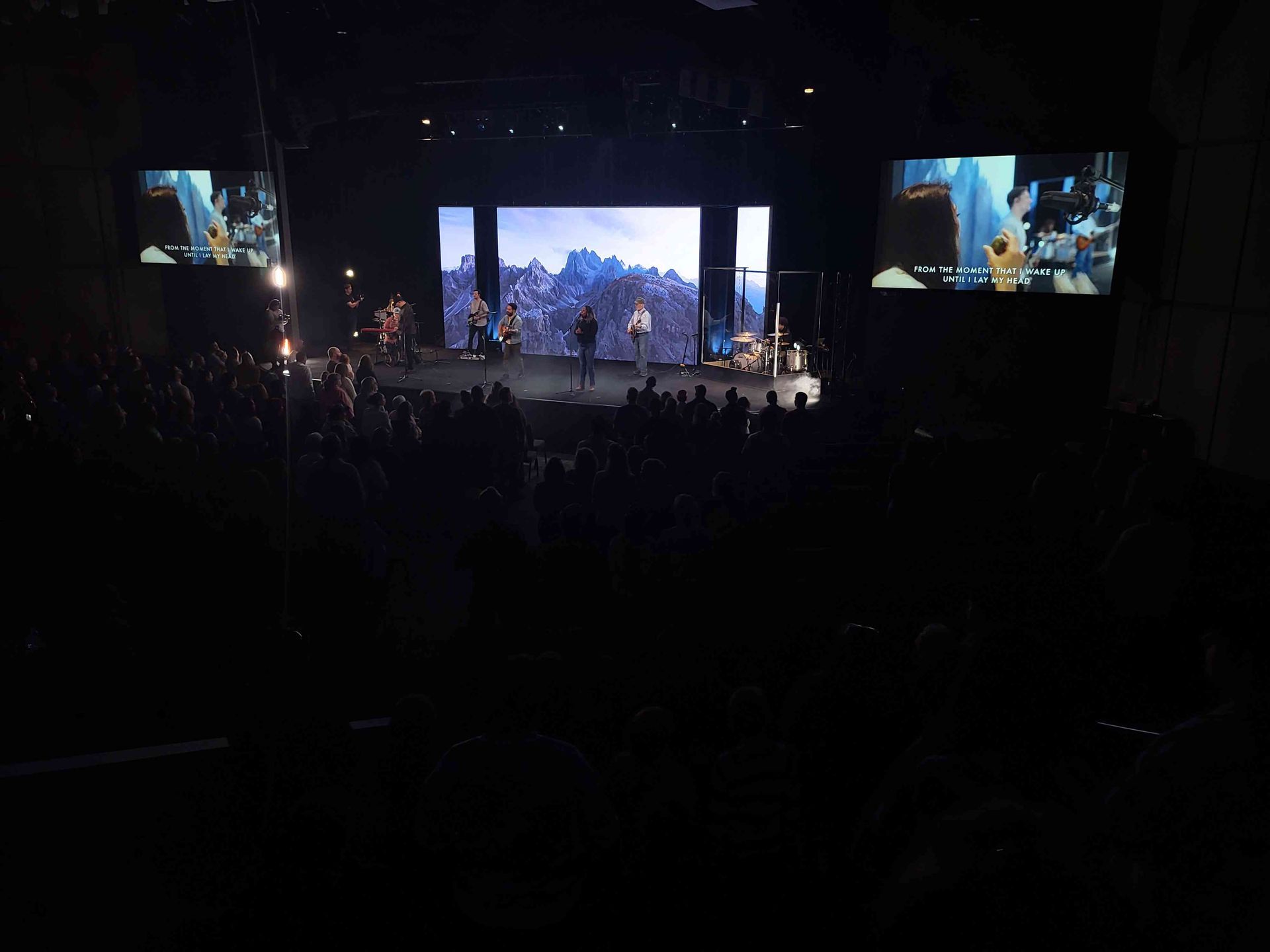 Audience watches a band perform on stage, screens display images, in a dark auditorium.