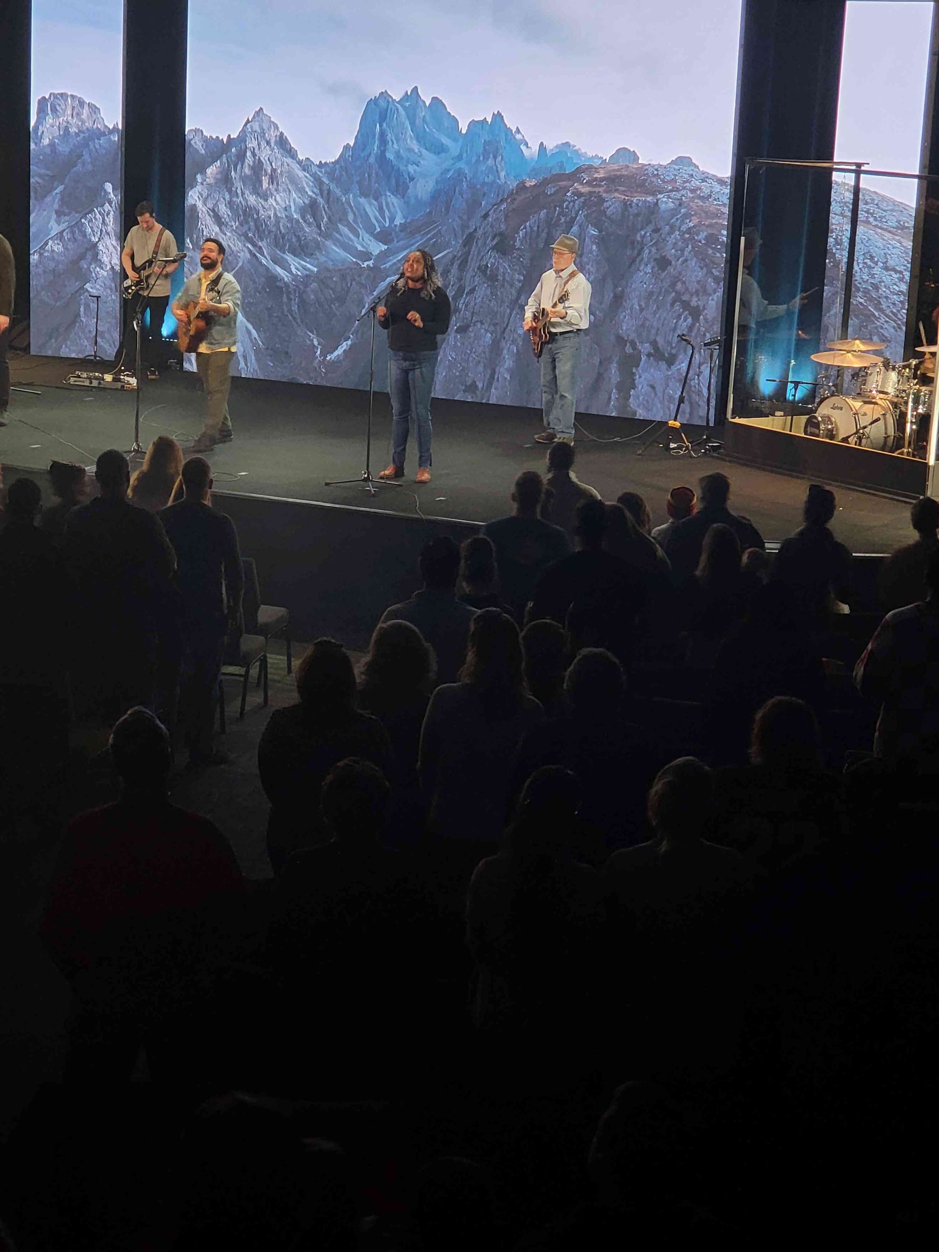 Band performing on stage with mountainous backdrop, audience watching in low light.