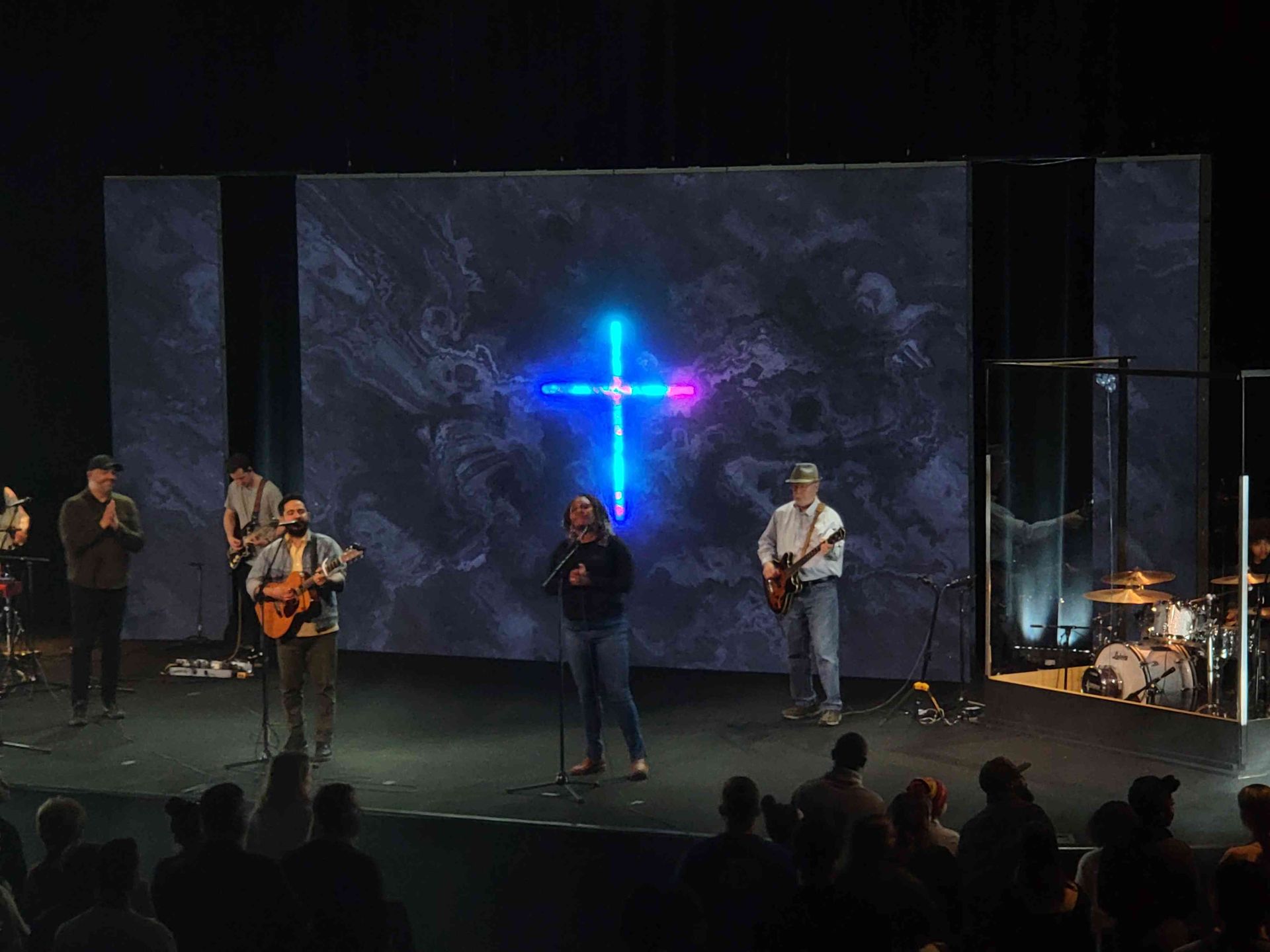 Worship band on stage with cross backdrop. Singer center, guitarists, drummer, and audience present.