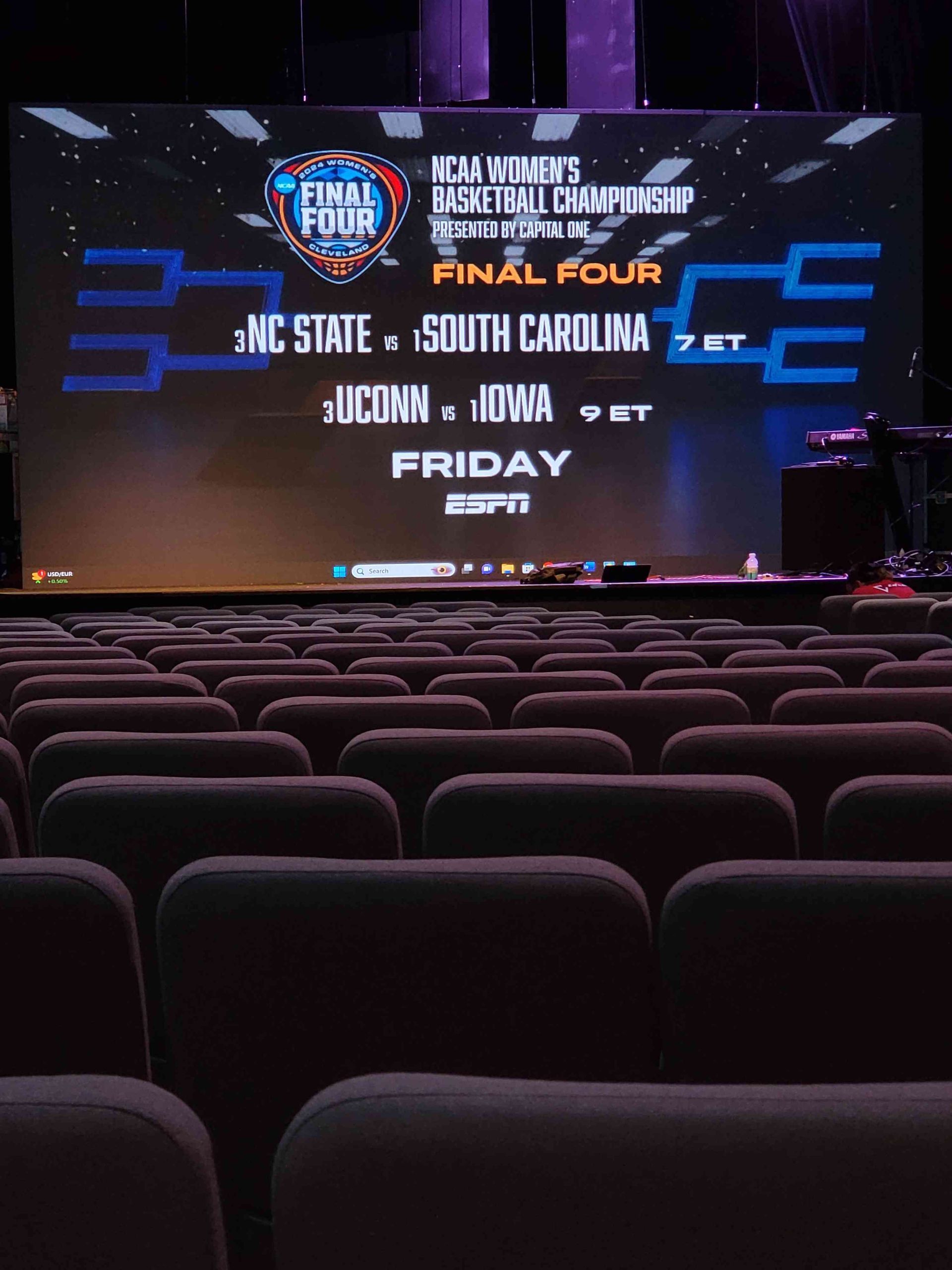 NCAA Women's Basketball Final Four bracket displayed on a large screen in an auditorium. Teams include NC State, South Carolina, UConn, Iowa.