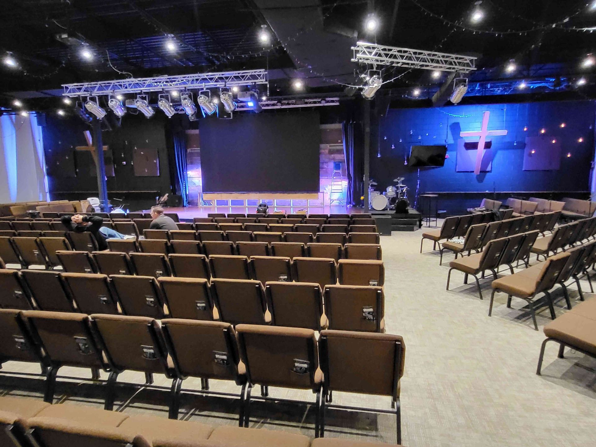 Rows of brown chairs face a stage with a large screen and cross, church interior.