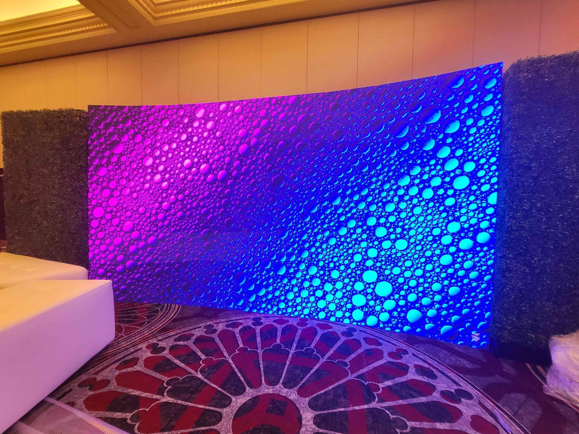 Curved LED screen with a purple and blue bubbly pattern, flanked by green walls. On a patterned rug.