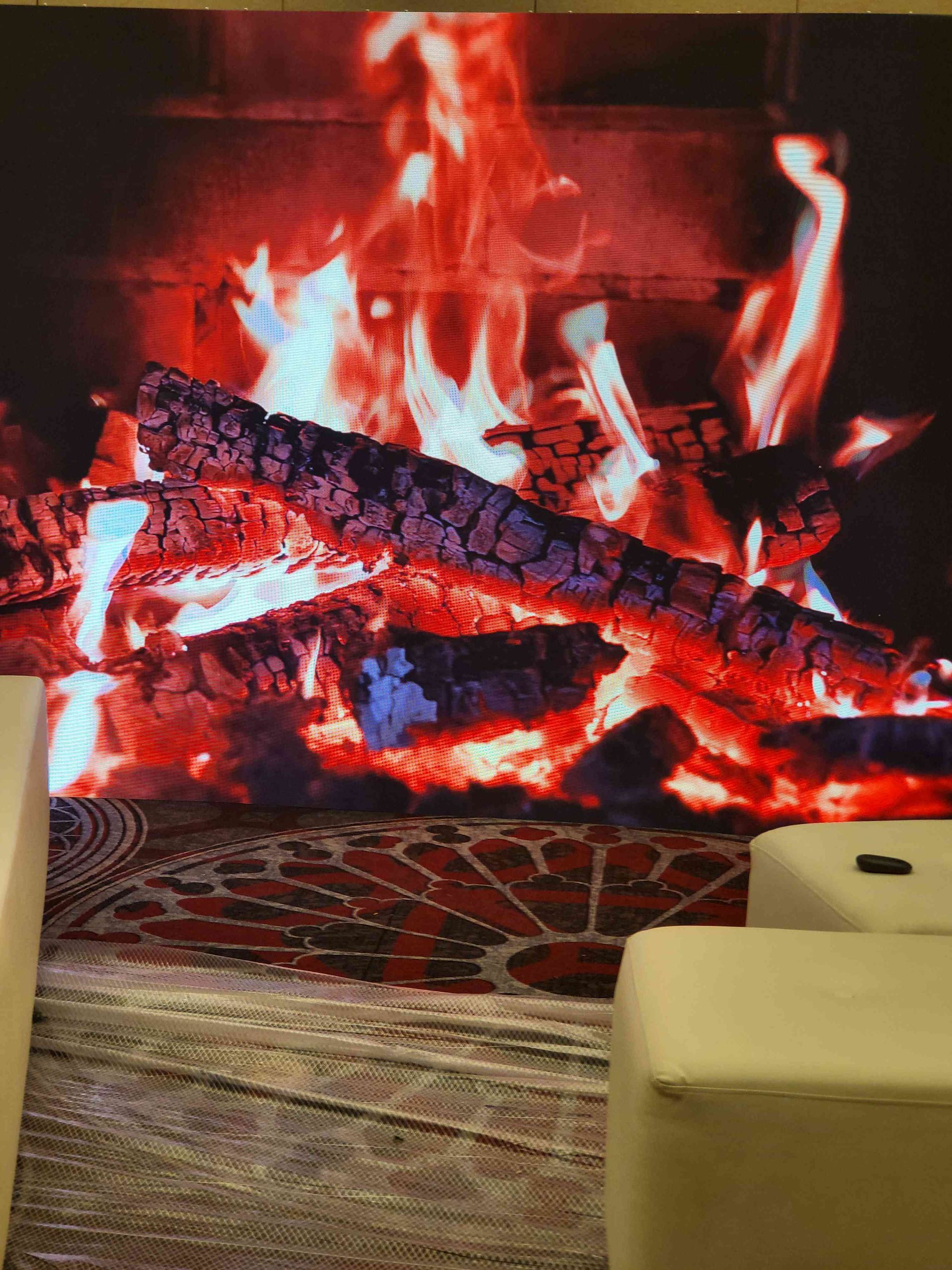 A large screen displaying a roaring fireplace with red and orange flames, wood, and a patterned rug.
