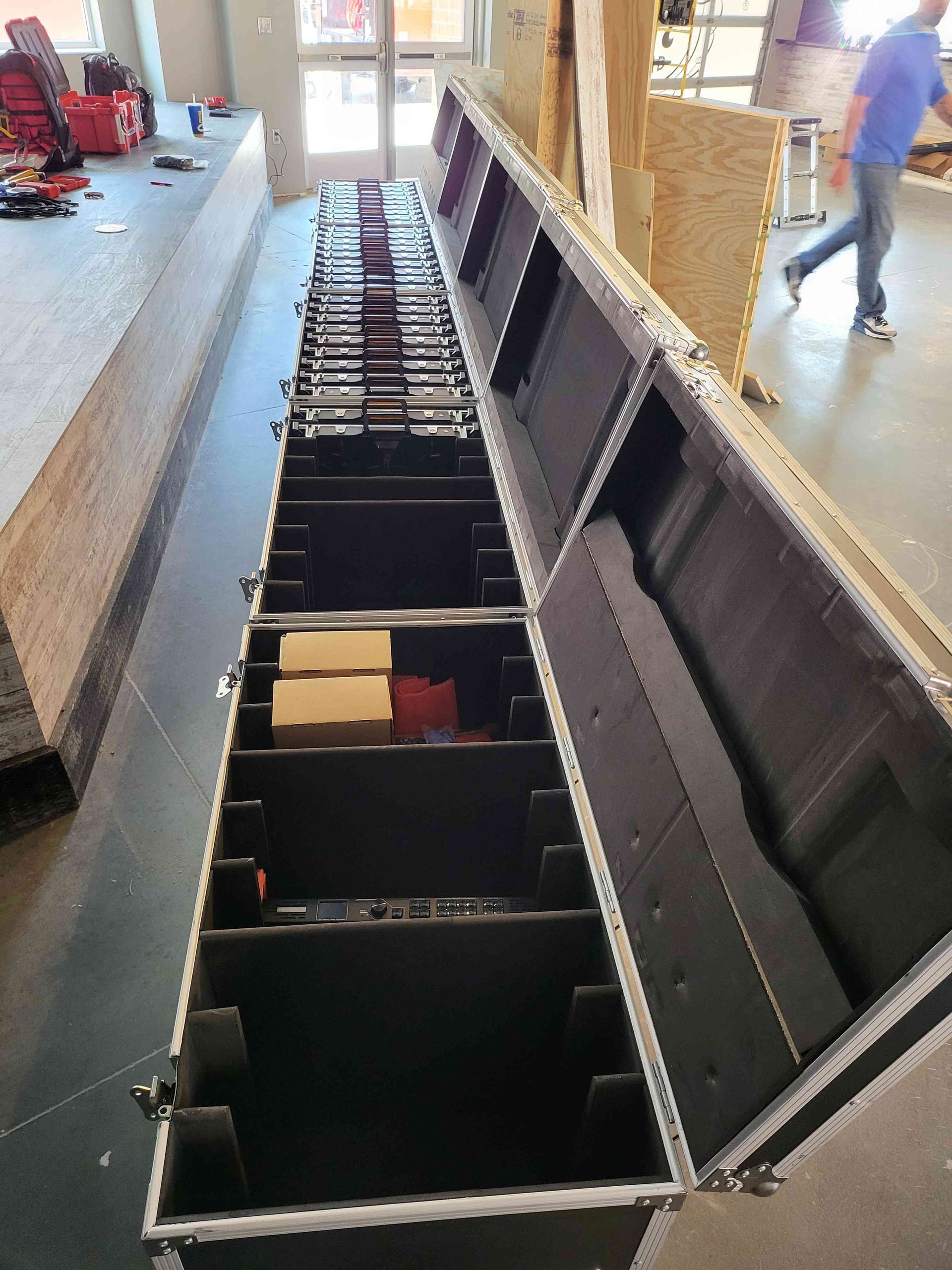 Open, black equipment case filled with gear, partially open in a construction area, with a person walking by.