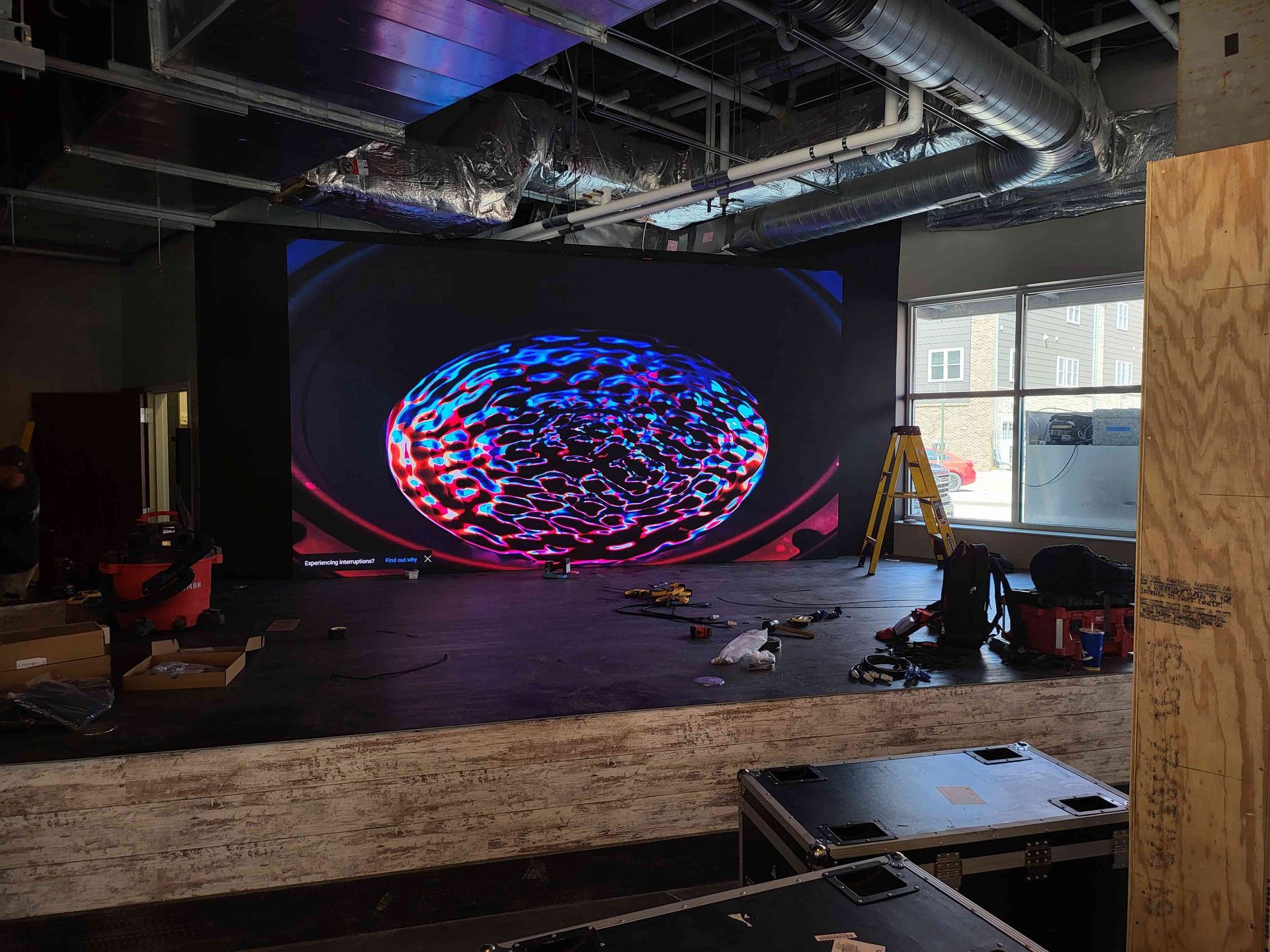Large LED screen displays colorful abstract shape in a venue under construction; tools and equipment scattered.