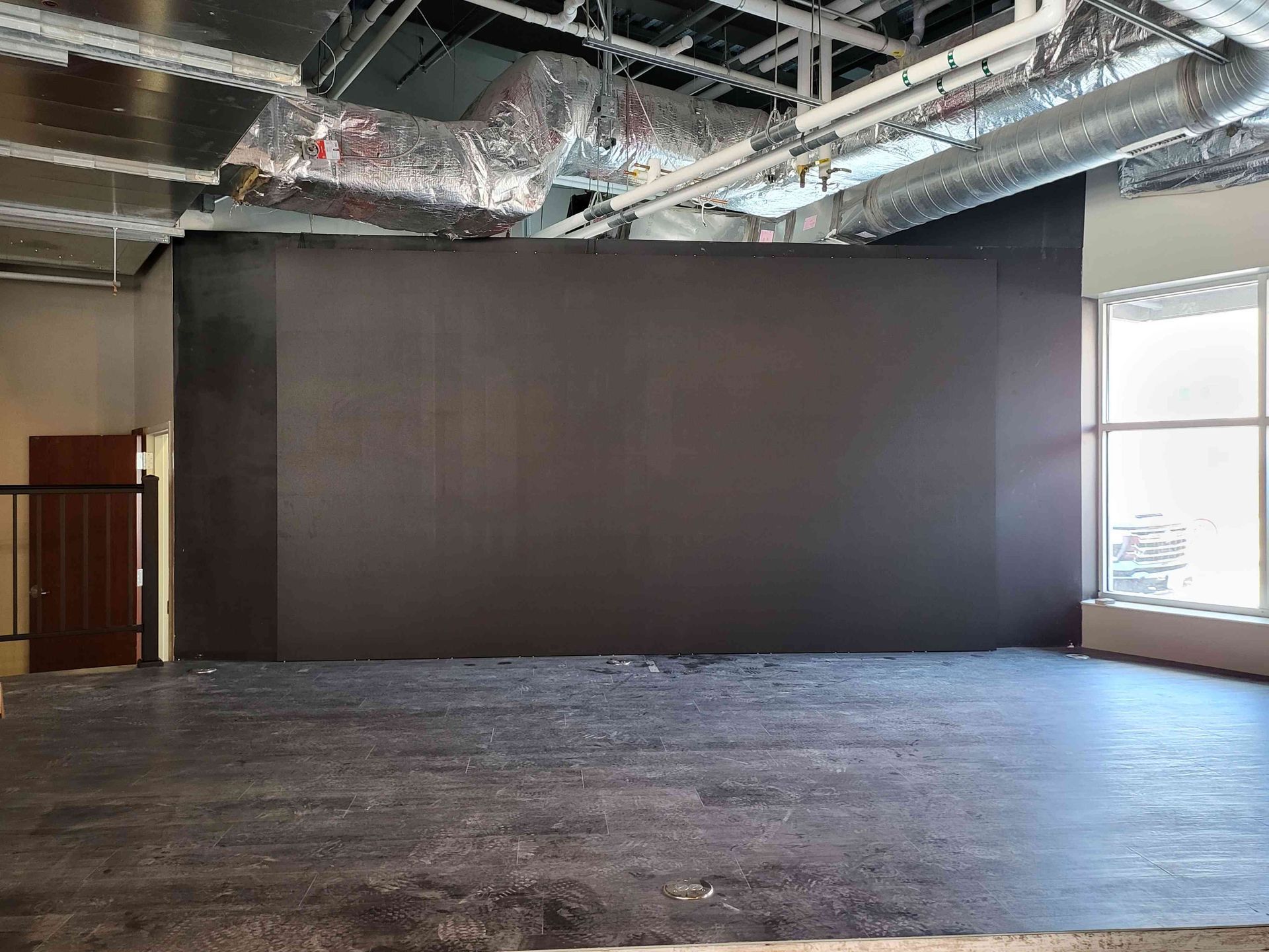 Empty stage with a large dark screen, a door on the left, and a window on the right. The floor is dark.