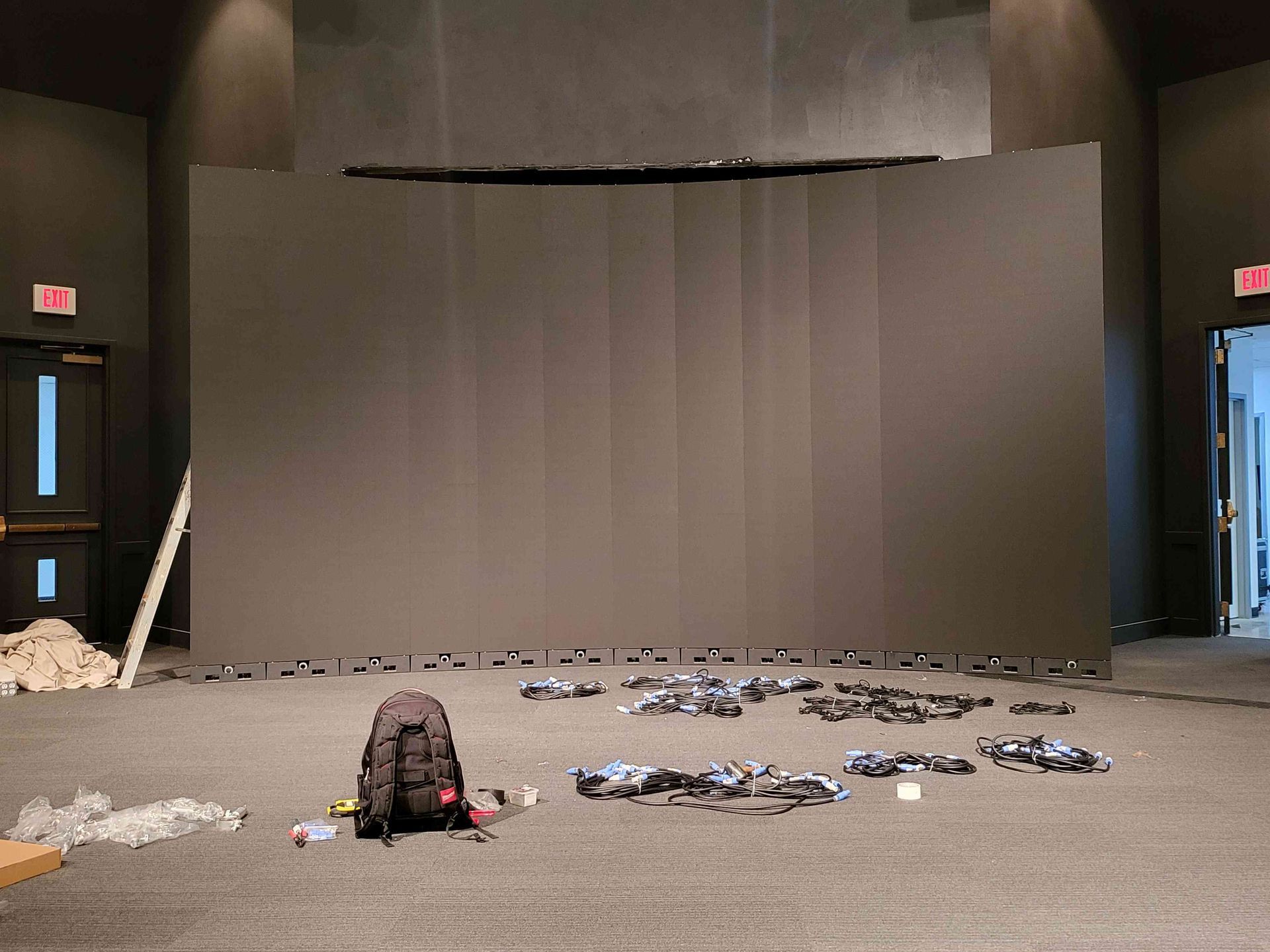 Large, curved black screen dominates a room; various tools and equipment scattered on the floor in front of it.