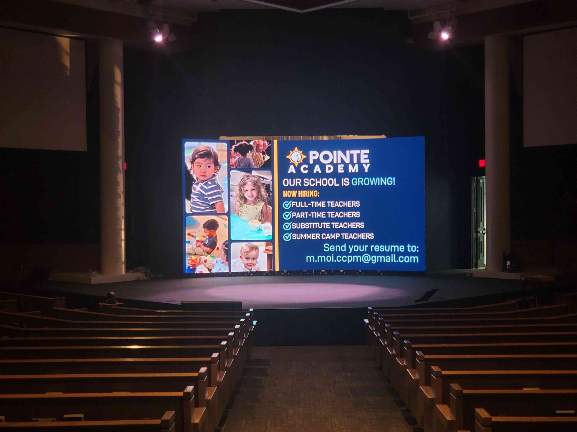 Interior of an auditorium. LED screen displays an advertisement for Pointe Academy. Rows of wooden pews face the stage.