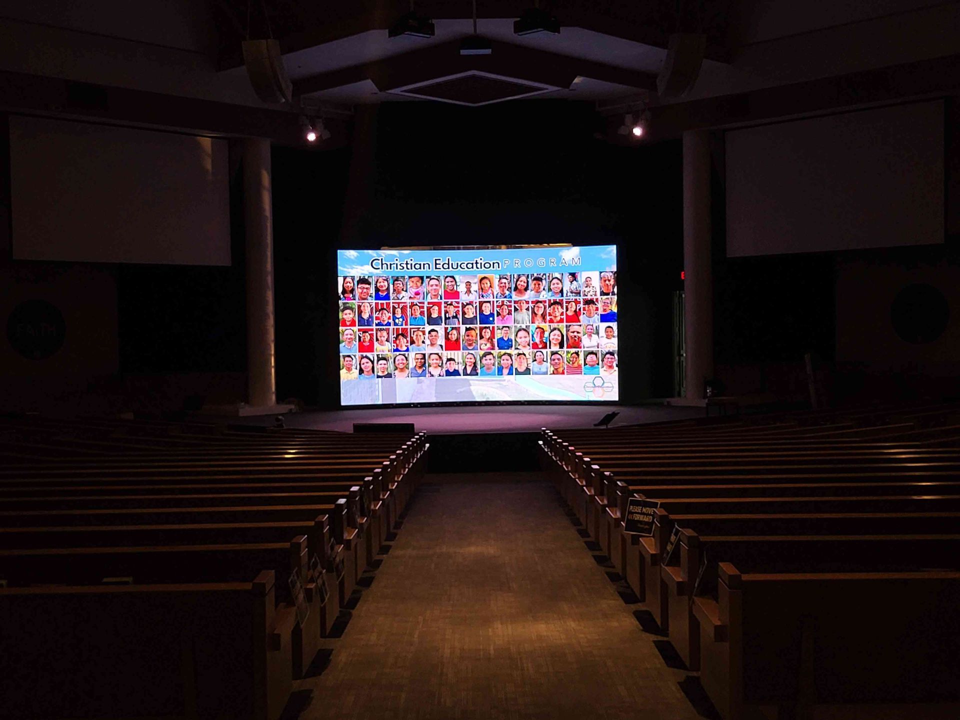 Interior of a church; large screen displays a collage of faces above a stage. Empty pews face the screen.
