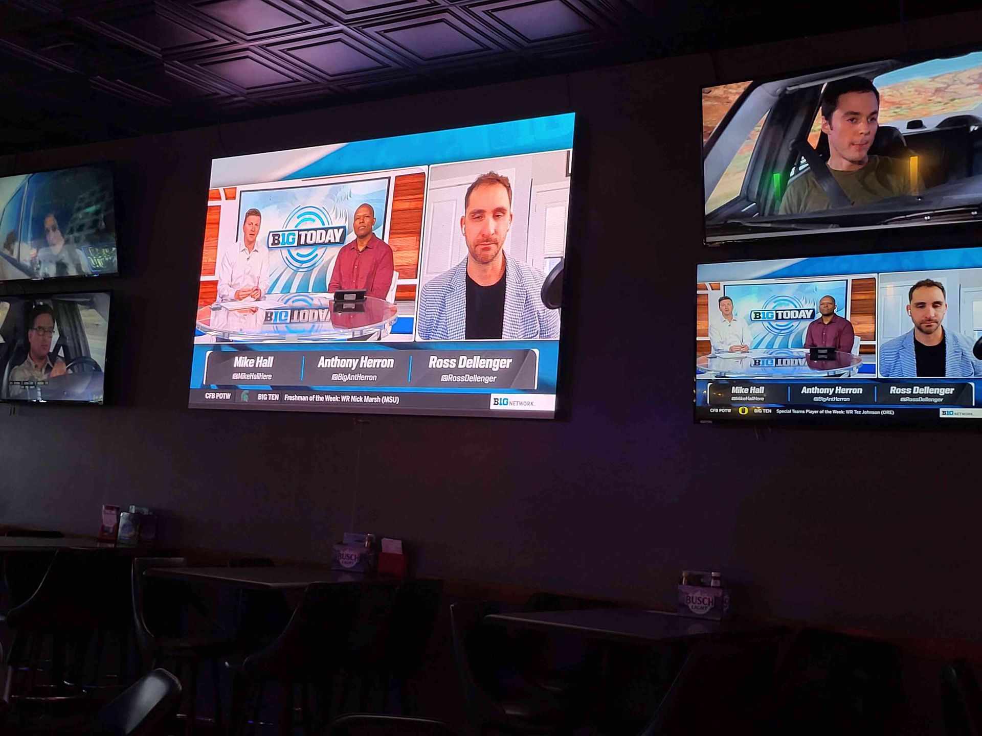 TVs on a wall in a bar show a sports talk show, plus a car driving scene. Dark room with tables.