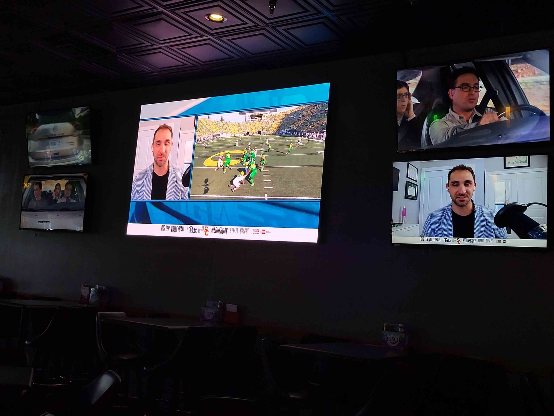 Dark bar with multiple TVs showing a man in a striped shirt. One shows a football game.